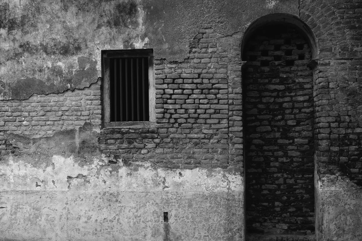 Un muro di mattoni antico con un’apertura ad arco tamponata da una muratura più recente, il cui contorno resta visibile, e accanto una piccola finestra con inferriata; fotografia in bianco e nero, nessuna persona.