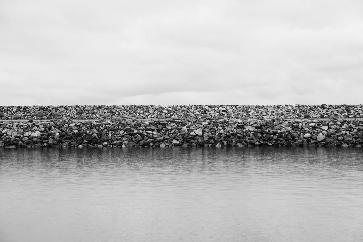 A breakwater of large rock armour lined up along the shore of a body of water, overcast sky, black-and-white photograph