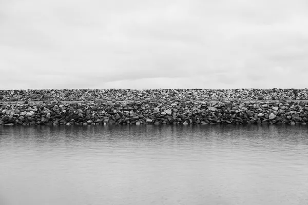 A breakwater of large rock armour lined up along the shore of a body of water, overcast sky, black-and-white photograph