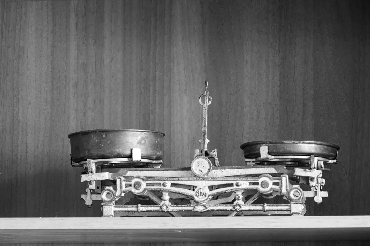 Bilancia a due piatti in ottone e ferro su un piano di legno davanti a una parete di legno, con il piatto di sinistra più basso dell’altro, fotografia in bianco e nero