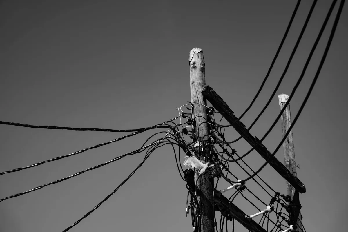 Wooden pole with several bundles of aerial cables tangled and under tension towards neighbouring poles, even sky, in black and white