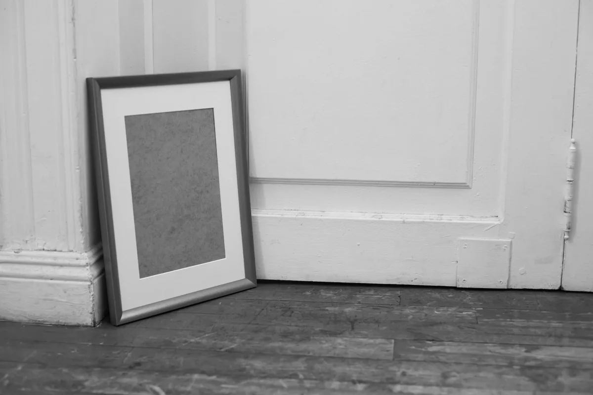 Cornice di metallo appoggiata a una parete bianca, con il passe-partout bianco e il cartone grezzo del fondo al posto della fotografia, immagine in bianco e nero