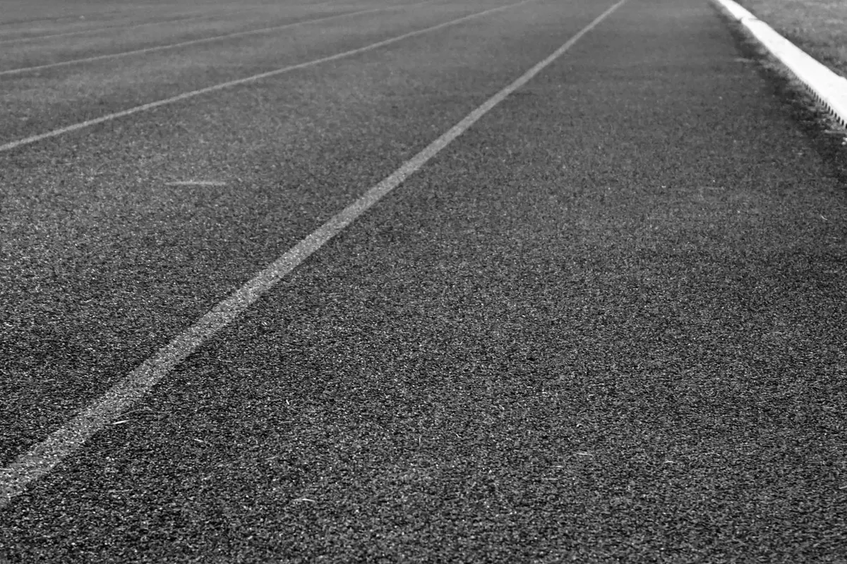 Pista di atletica in bianco e nero ripresa radente da terra, corsie bianche tracciate che corrono in prospettiva verso il fondo, manto granuloso in primo piano, cordolo di scolo chiaro, striscia d’erba sul lato destro