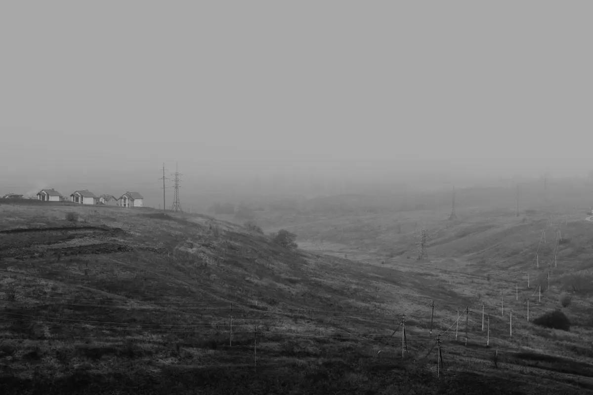 Houses on a ridge at the edge of a valley in fog, with poles and line pylons vanishing into the white