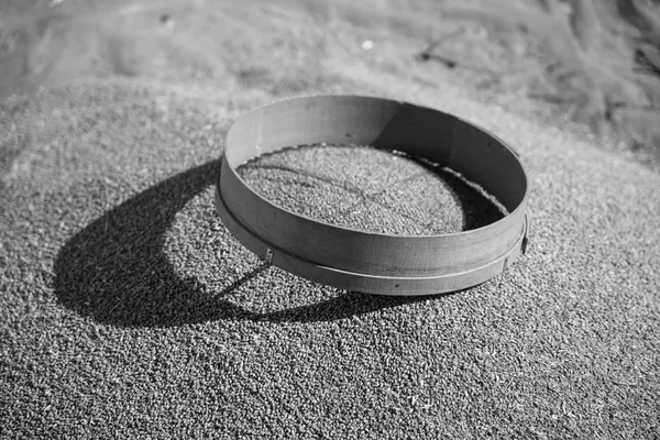 A circular wooden-rimmed riddle resting on a heap of wheat grains in the sun, its shadow cast across the heap, black and white photograph