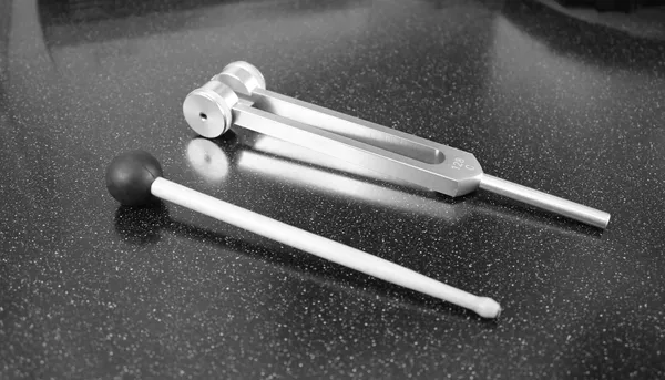 Metal tuning fork with adjustable weights and a wooden mallet lying on a dark speckled worktop, in black and white