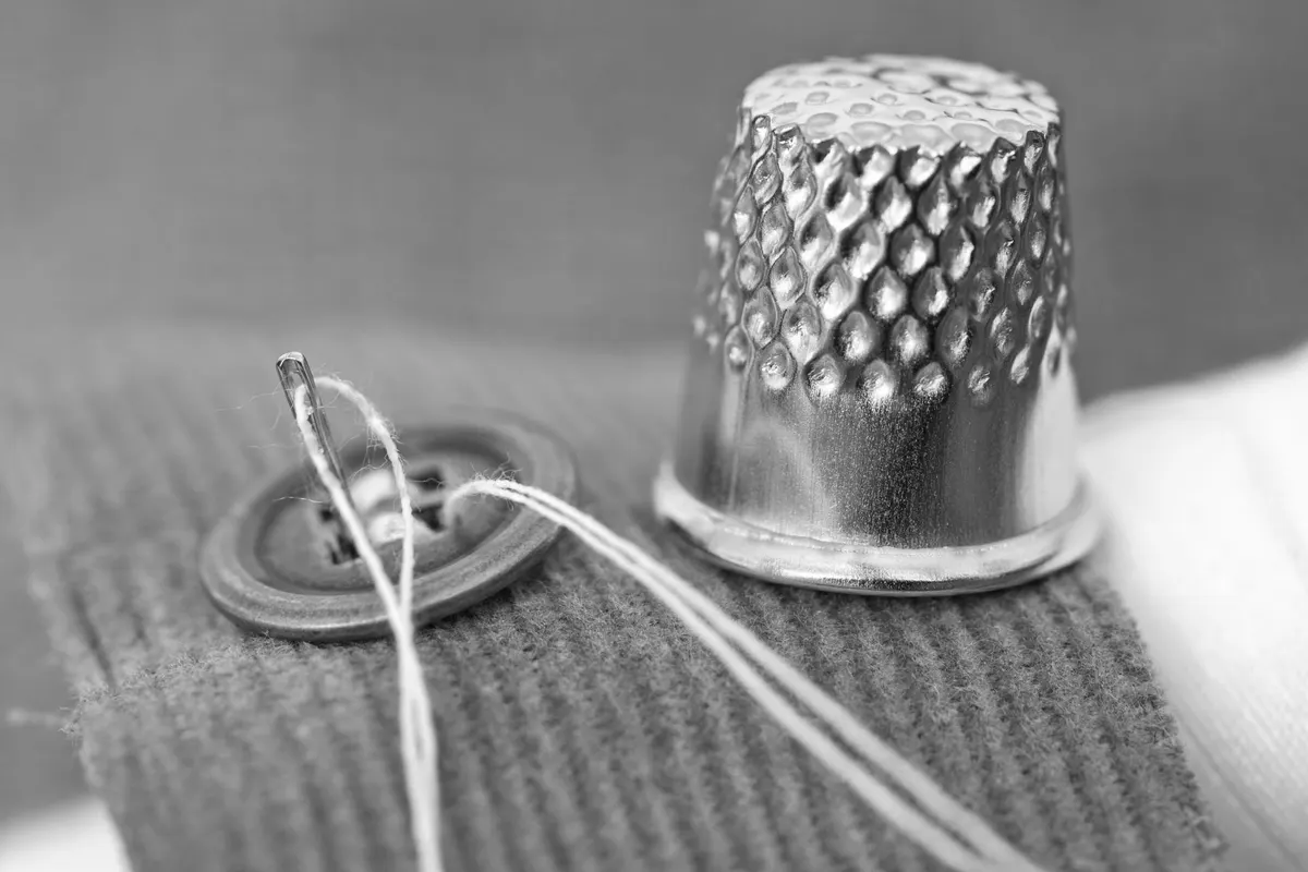 A metal sewing thimble, a threaded needle and a button on a scrap of ribbed fabric, black and white macro photograph