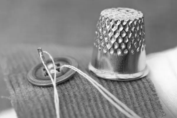 A metal sewing thimble, a threaded needle and a button on a scrap of ribbed fabric, black and white macro photograph
