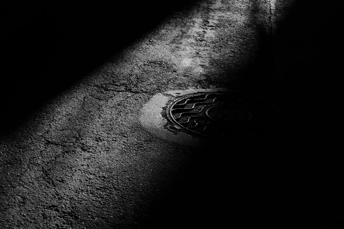 Un fascio di luce diagonale taglia l’asfalto bagnato fino a un chiusino a raso, di notte, fotografia in bianco e nero