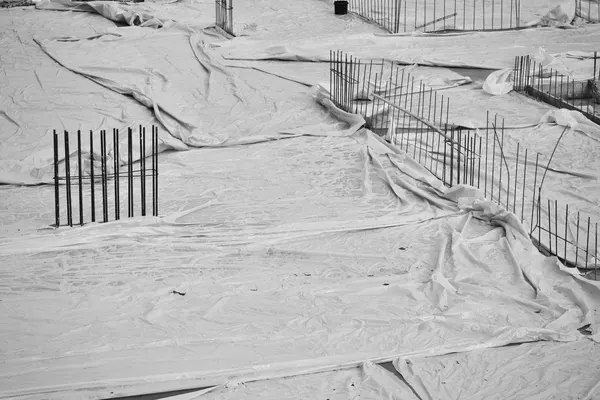 Reinforcement bars protruding from a foundation slab covered with polythene sheeting, in black and white