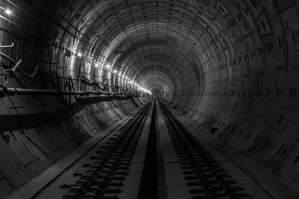 Railway tunnel under construction, lit by a row of lights along the curved wall, with cables and conduits fixed alongside the tracks, in black and white