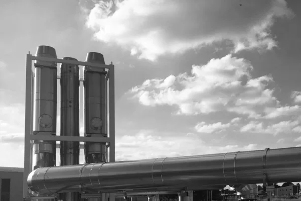Exhaust stacks and a large lagged duct on a building’s rooftop plant, against the sky, black and white photograph