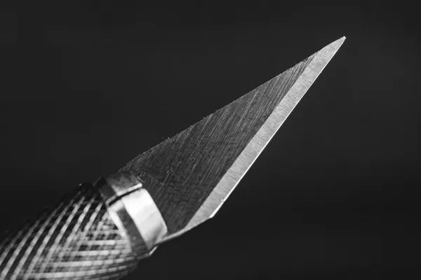 The tip of a thin blade with its edge cut at a slant, shot in macro against a dark background, in black and white