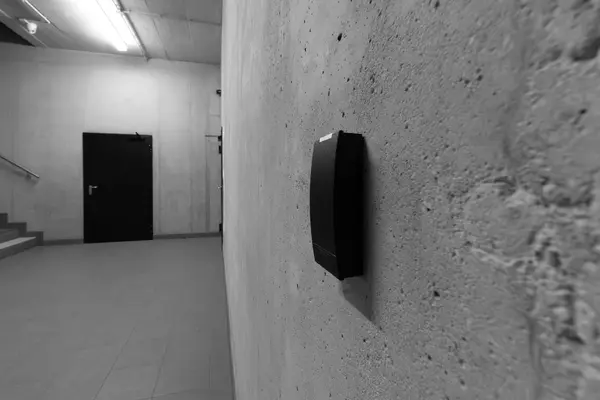 Black badge reader on an exposed concrete wall, technical corridor with a closed metal door in the background, black and white photograph