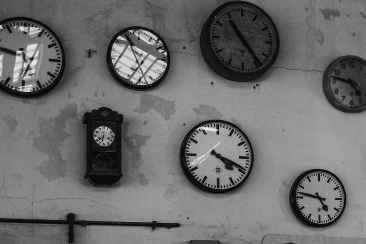 A peeling wall with about a dozen wall clocks of different styles, each showing a different time, black-and-white photograph