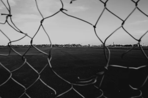 A chain-link fence with a torn wire strand in the foreground, seen through the mesh onto a paved yard with buildings in the distance, black-and-white photograph