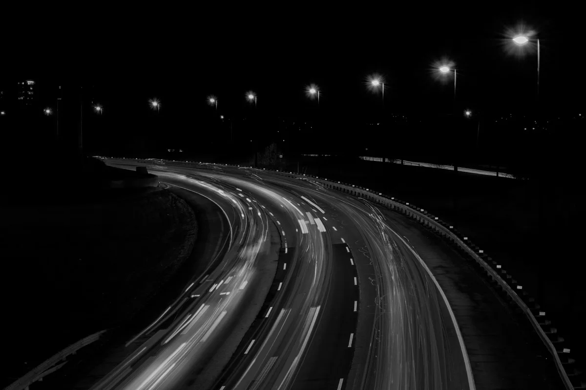 Scie luminose di traffico notturno su una carreggiata curva, in bianco e nero, con lampioni accesi sullo sfondo