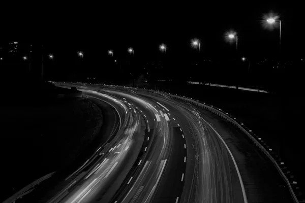 Scie luminose di traffico notturno su una carreggiata curva, in bianco e nero, con lampioni accesi sullo sfondo