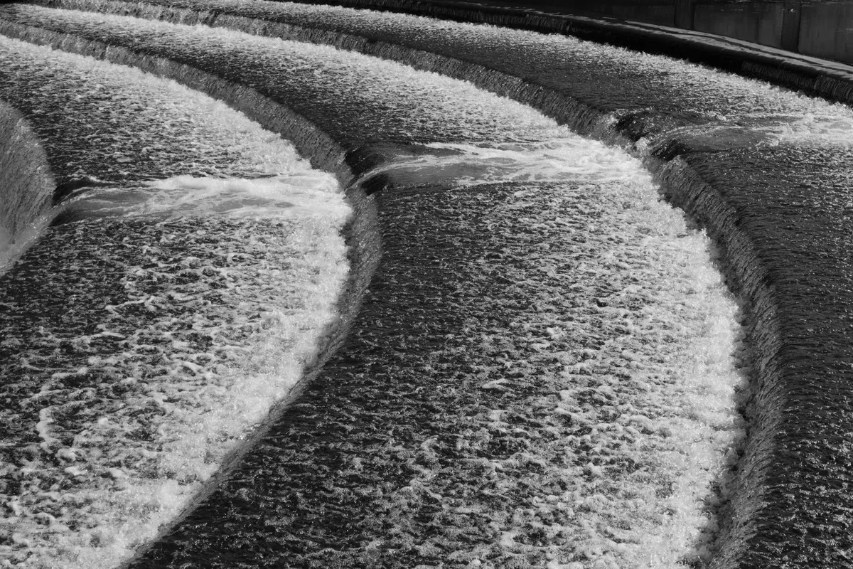 Water spilling over two curved concrete sills of a weir, foam along the crest, black and white photograph