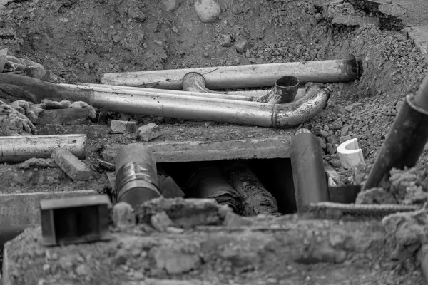 Pipes and conduits of different materials and diameters piled together in an excavation, among soil and rubble, black-and-white photograph
