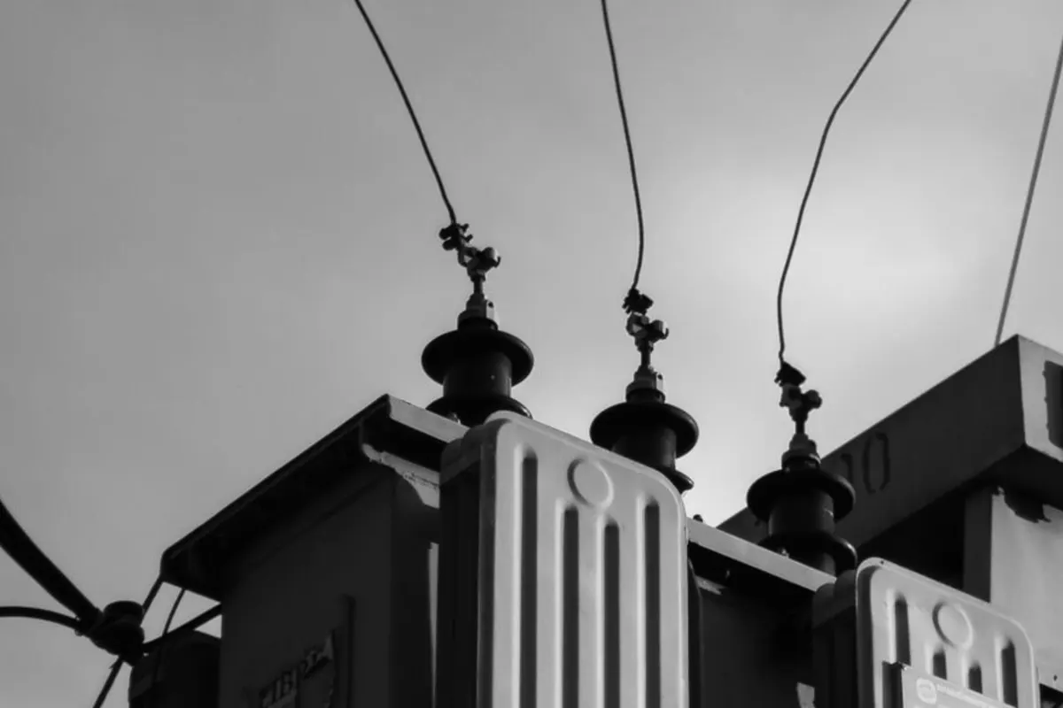 Three dark ceramic bushing insulators on top of an electrical transformer, thin wires rising into an overcast sky, pale ridged cooling fins in the foreground, black and white photograph