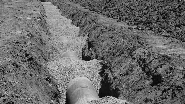 A construction trench with three corrugated grey ducts laid on a gravel bed, disturbed red earth to either side, in black and white
