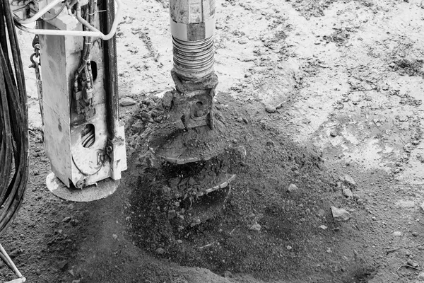 The helical head of an auger drill breaking through loose soil, beside the mast of the rig and its hydraulic hoses, close-up black-and-white photograph